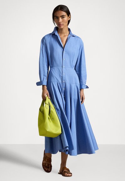 Modra srajčna obleka z gumbi spredaj, ovratnikom, dolgimi rokavi in rahlo padajočo krilno linijo. Kombinirana s svetlo zeleno torbico in rjavimi sandali.