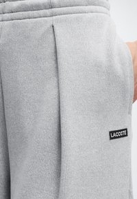 Gros plan sur un pantalon en maille gris clair avec un pli sur le côté et un patch logo Lacoste rectangulaire noir près de la poche sur la hanche droite.