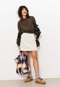 Chemise boutonnée à rayures brunes, jupe en velours côtelé crème, chaussettes grises, chaussures beiges à lacets, et un sac fourre-tout à carreaux bleus avec des accents orange.