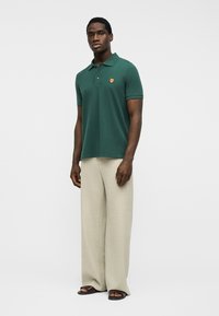 Man wearing a dark green polo shirt with an embroidered fox patch, beige wide-leg pants, and dark open-toe sandals on a plain background.