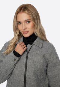 Grau gestreifte Jacke mit Kragen und einem Frontreißverschluss. Strukturierter Stoff mit lockerem Sitz und schwarzem Rollkragenpullover darunter.