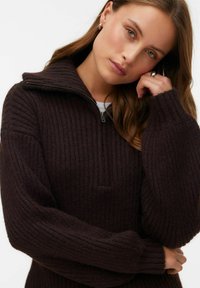 Mladá žena se světle hnědými vlasy, oblečená ve tmavě hnědém žebrovaném svetru s límečkem na zip do čtvrtiny, opírající si hlavu o ruku.