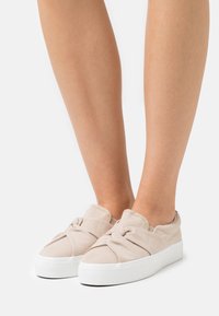 Beige canvas slip-on shoes with a knotted detail on the front, featuring a thick white sole and a textured rubber bottom.