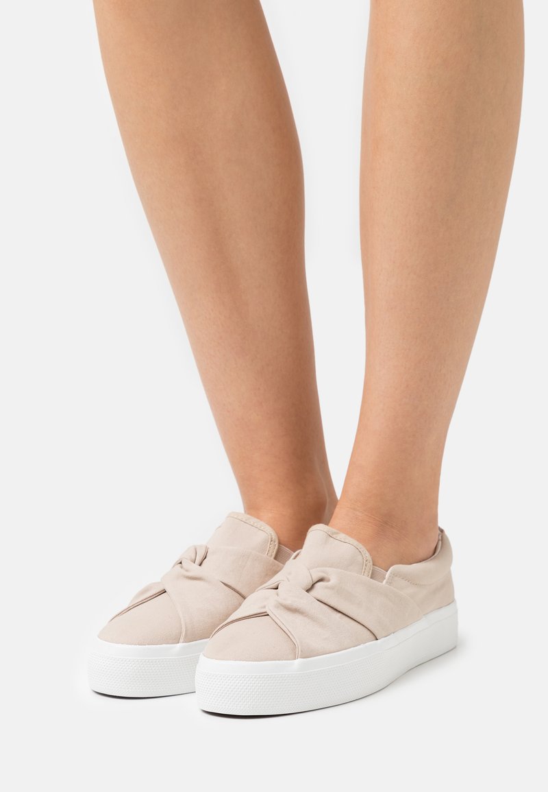 Beige canvas slip-on shoes with a knotted detail on the front, featuring a thick white sole and a textured rubber bottom.