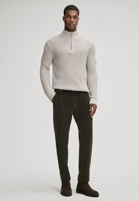 Pull en maille beige avec col haut et demi-zip, associé à un pantalon en velours côtelé vert foncé et des bottines en daim marron.