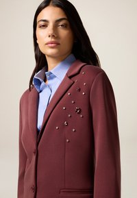 Blazer bordeaux con un design aderente, caratterizzato da gemme decorative su un lato e una camicia azzurra con colletto sotto. Texture liscia.