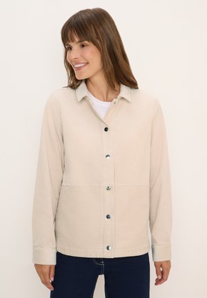 Femme souriante, portant une veste en côtes beige à boutons sur un haut blanc, associée à un jean bleu foncé, se tenant contre un fond clair.