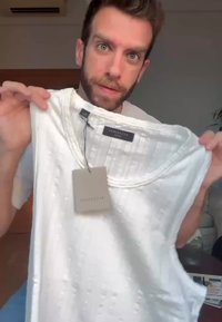 Man met baard houdt een witte mouwloze gebreide blouse met merketiket omhoog in een binnensetting.