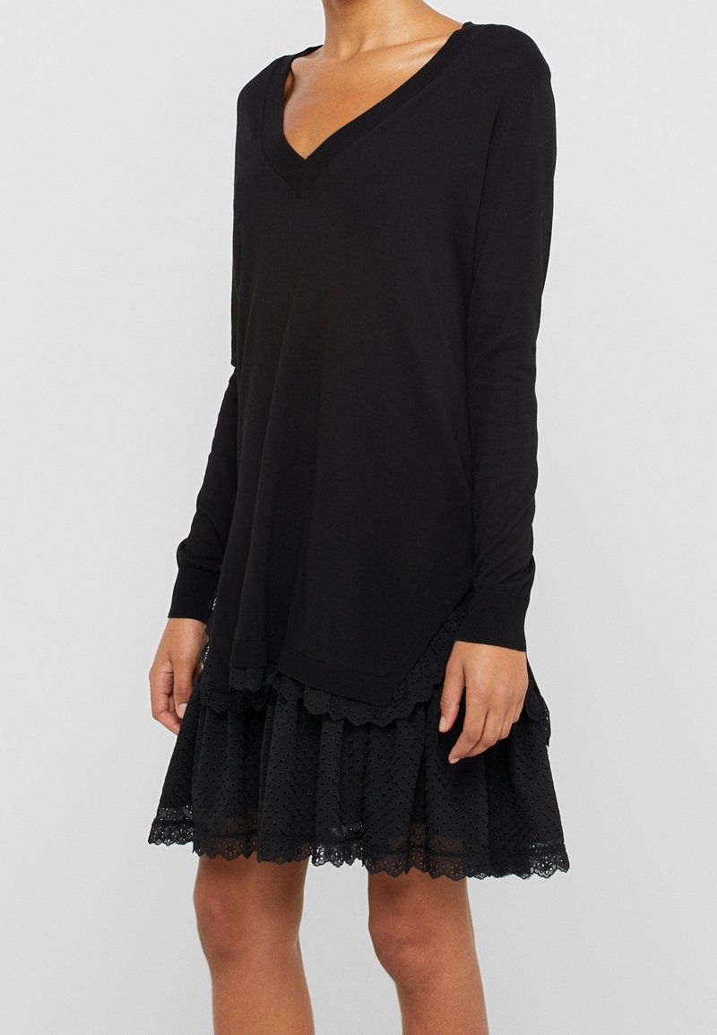 Femme portant un haut noir à manches longues en V sur une jupe en dentelle noire aux bords festonnés sur un fond clair uni.