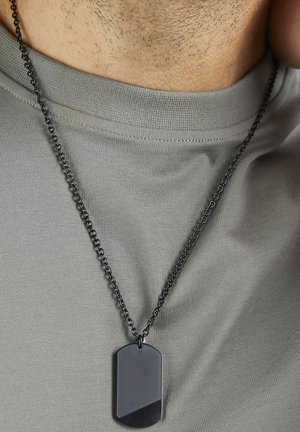 Schwarzes Hundemarkenanhänger an einer schwarzen Kette, getragen über einem hellgrauen T-Shirt. Der Anhänger hat eine glatte, flache Oberfläche mit abgerundeten Kanten.