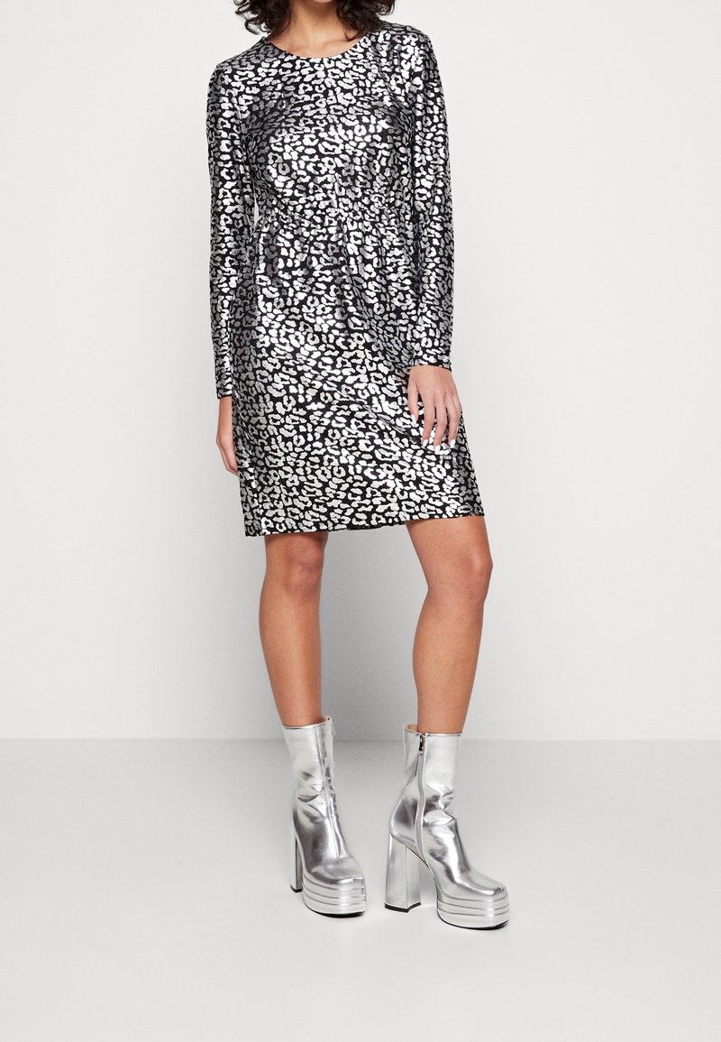 Femme portant une robe à manches longues argentée et noire à motifs, avec des bottines plateformes argentées brillantes, debout devant un fond uni.