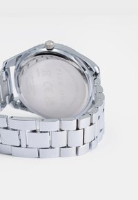 Ceas de mână din oțel inoxidabil cu un finisaj argintiu lustruit, brățară din metal cu verigi și carcasă rotundă, netedă. Brandul gravat este vizibil pe spate.