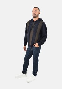 Felpa con cappuccio a maglia blu navy e marrone, con parte frontale testurizzata, abbinata a jeans scuri e sneaker bianche. Il design presenta un cappuccio e polsini a coste.