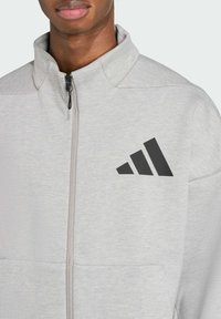 Svetlo šedá bunda na zips vyrobená z textúrovanej tkaniny, s čiernym logom Adidas a tromi diagonálnymi pruhmi na ľavom hrudníku a dvoma prednými vreckami.