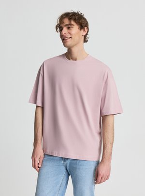 Giovane uomo sorridente, che indossa una maglietta rosa chiaro larga e jeans azzurri, in piedi davanti a uno sfondo chiaro uniforme.