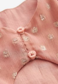 Tissu pêche avec une finition texturée, présentant de petits motifs dorés et deux boutons roses au niveau du col, chacun arborant un design floral.