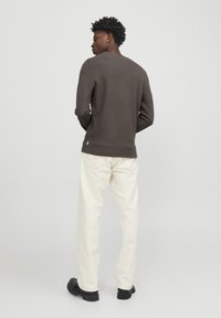 Camisa de manga larga marrón y texturizada con cuello redondo, combinada con pantalones color crema y zapatos negros, vista desde la parte de atrás.