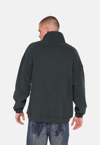Sweatshirt oversize vert foncé avec un col haut, texture douce, épaules tombantes et coutures détaillées au dos, porté avec un jean bleu clair.