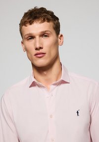 Camicia di cotone rosa chiaro con colletto classico, chiusura a bottoni e logo blu navy sul petto. Tessuto morbido e vestibilità sagomata.