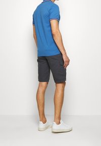Uomo che indossa una polo blu, pantaloni cargo grigi scuri e sneakers bianche, in piedi contro uno sfondo bianco semplice.