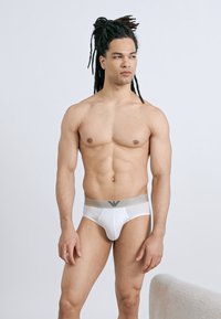 Μυώδης άνδρας με dreadlocks φοράει λευκά εσώρουχα με γκρι ζώνη στη μέση, κοιτάζοντας προς τα πλάγια σε απλό φόντο.