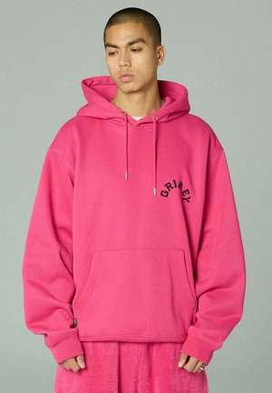 Junge Person mit kurz geschorenem Haar, trägt einen übergroßen, leuchtend pinken Hoodie mit dem Schriftzug "GRIMEY" auf der Brust, steht vor einem schlichten grauen Hintergrund.