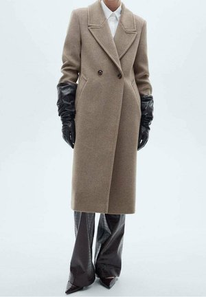 Femme portant un long manteau beige à double boutonnage, des gants en cuir noirs, un pantalon large texturé noir et des chaussures noires à bout pointu.