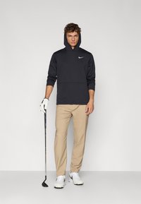 Dark grey hoodie with a kangaroo pocket and white Nike logo, paired with beige pants, white sneakers, and holding a golf club.