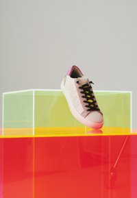 Baskets blanches avec lacets noirs et talon rose reposant sur des cubes d'exposition transparents jaunes et rouges sur un fond neutre.