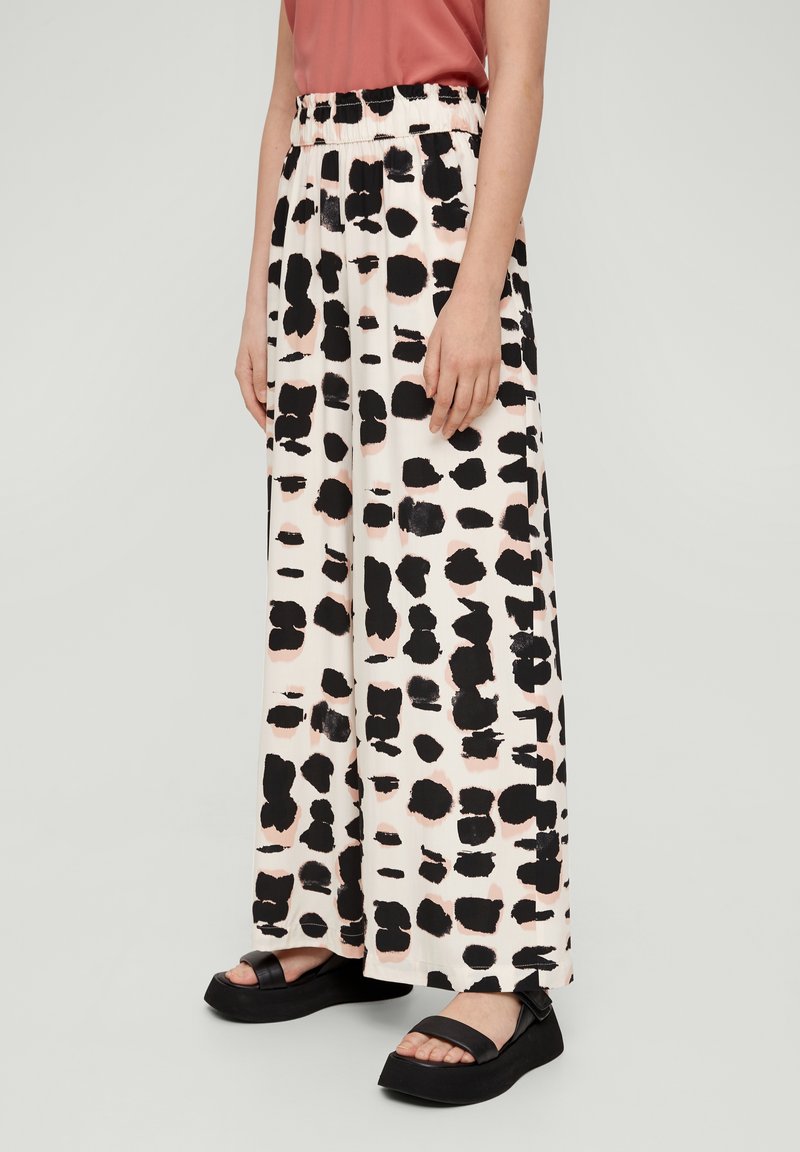 Femme portant un pantalon blanc à jambes larges avec des taches abstraites noires et des sandales compensées noires, debout devant un fond uni.