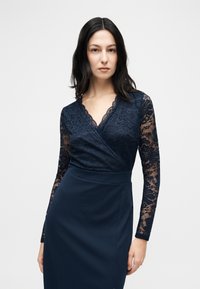 Γυναίκα με μακριά σκούρα μαλλιά, ντυμένη με φόρεμα σε ναυτικό μπλε χρώμα, με δαντελένια μακριά μανίκια και V-λαιμόκοψη, στέκεται μπροστά σε ένα απλό ανοιχτόχρωμο φόντο.