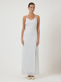 Γυναίκα φορεμένη με ένα άσπρο αμάνικο φόρεμα καλυμμένο με παγιέτες, με V-λαιμόκοψη, πλαϊνό άνοιγμα, και μπεζ ψηλές γόβες, να στέκεται μπροστά σε έναν απλό φόντο.