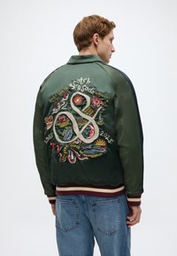 Πράσινο σατινέ μπουφάν τύπου bomber με λεπτομερή κεντήματα στην πλάτη, που περιλαμβάνουν φυτικά και ζωικά μοτίβα καθώς και κείμενο, με ριμπ μανσέτες και τελείωμα.