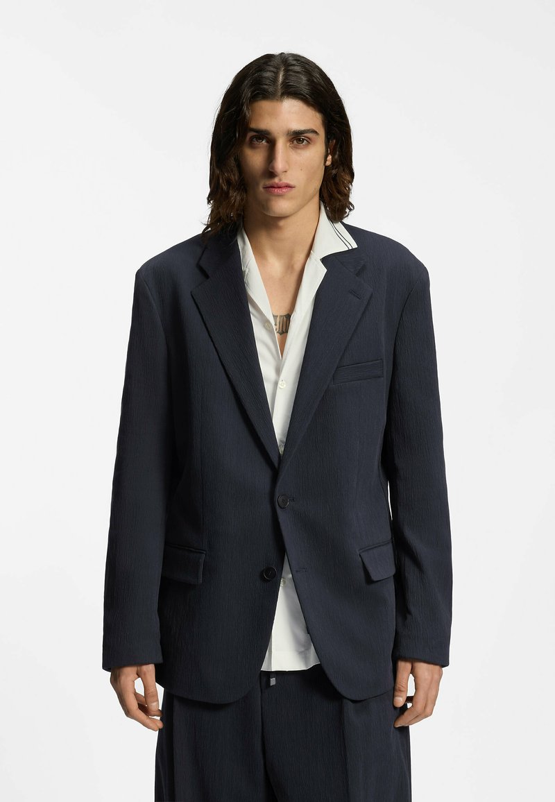 Jeune personne aux cheveux foncés mi-longs porte un blazer sombre oversize par-dessus une chemise blanche, debout devant un fond clair uni.