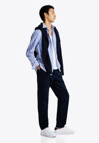 Conjunto masculino que incluye una camisa de botones a rayas azul y blanca, pantalones marinos y zapatillas blancas, con un suéter azul marino colgado sobre los hombros.