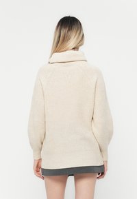 Cremfarbener Strickpullover mit hohem Kragen, langen Ärmeln und geripptem Muster, der eine lockere Passform und Seitenschlitze am unteren Saum aufweist.