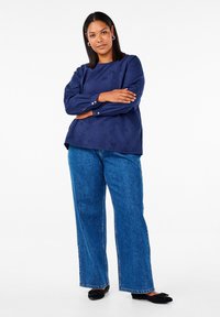 Mörkblå långärmad topp med en strukturerad mönster, parat med högmidjade vida blå jeans och svarta ballerina skor. Enkla guldknappar på ärmarna.
