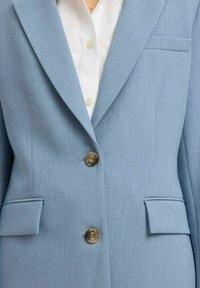 Blazer bleu clair avec deux boutons marron, poches à rabat, et chemise blanche en dessous, montré de la poitrine à la taille.