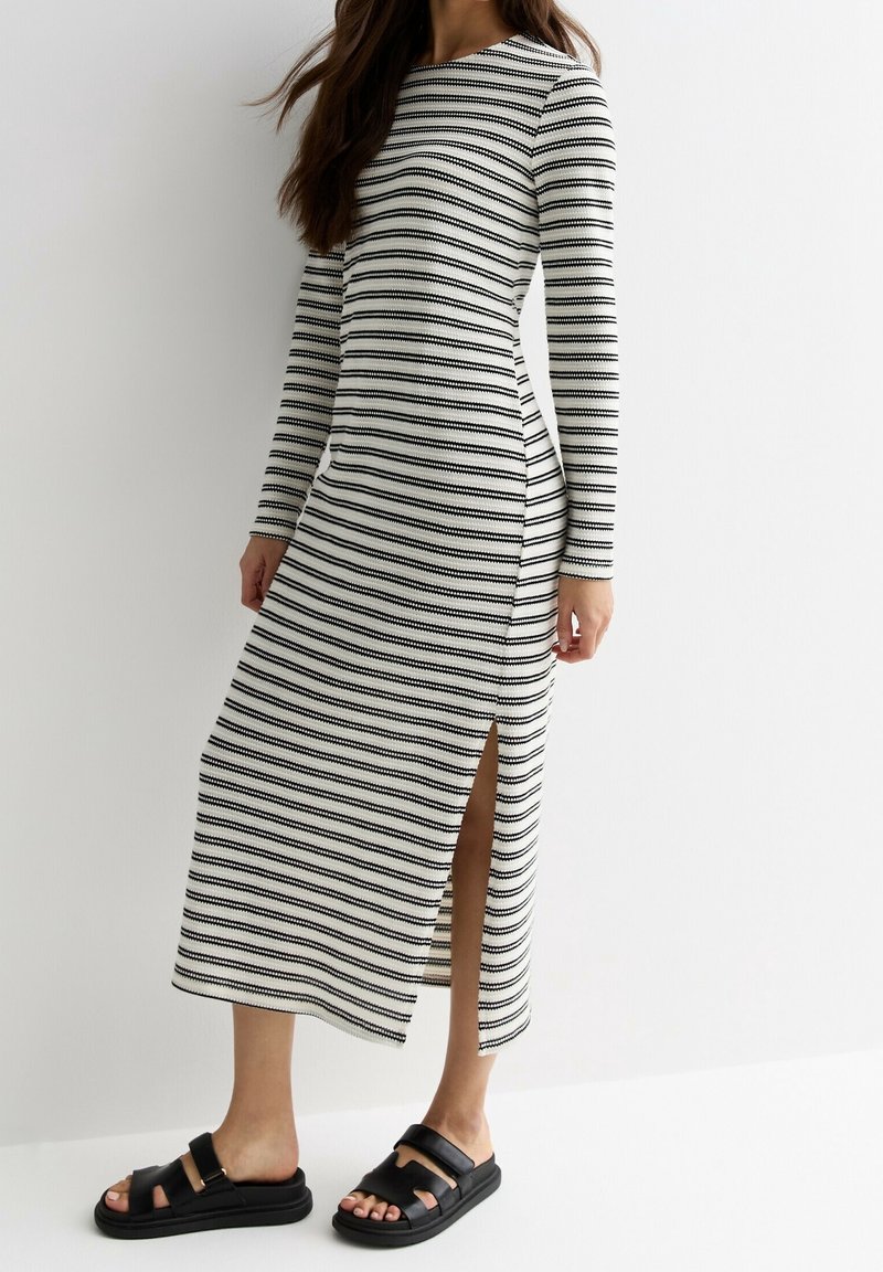 Femme portant une robe midi à manches longues rayée noir et blanc avec une fente latérale et des sandales noires à bouts ouverts, debout devant un fond blanc.