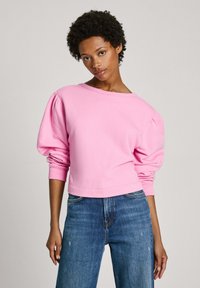 Rozā džemperis ar uzpūstām garajām piedurknēm, apļveida dekoltē un jostu. Kombinēts ar zilām džinsu biksēm, kas ir brīvā piegriezumā.