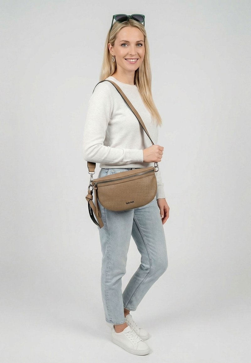 Vrouw draagt een witte trui, lichtblauwe spijkerbroek, witte sneakers, zonnebril op haar hoofd en een tan crossbodytas met zwarte banddetails.