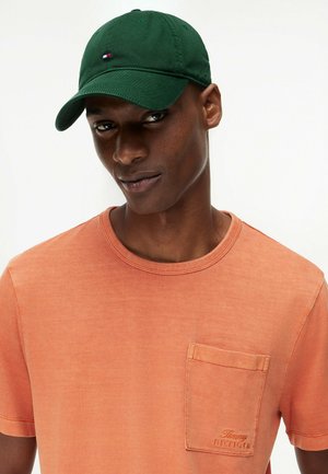 Man wearing a dark green cap and faded orange Tommy Hilfiger T-shirt with chest pocket, looking slightly sideways against a white background.