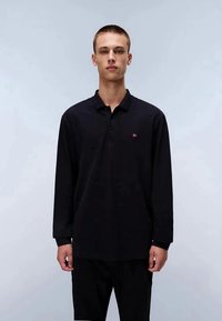 Jovem em pé, vestindo uma polo preta de mangas longas com um pequeno logo vermelho e branco no lado esquerdo do peito, fundo simples.