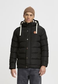 Doudoune noire avec un design matelassé, cordons de serrage crème, boutons en bois et un patch logo en cuir, avec une capuche et des poches latérales.