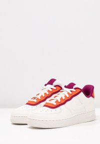 Sneaker in pelle bianca con accenti arancioni e viola, caratterizzati da una superficie liscia, punta perforata e suola in gomma bianca.