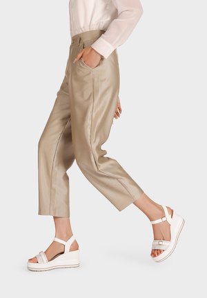 Pantaloni beige texturizzati con pieghe e tasche laterali abbinati a sandali con zeppa bianchi con cinghie alla caviglia regolabili e dettagli decorati.