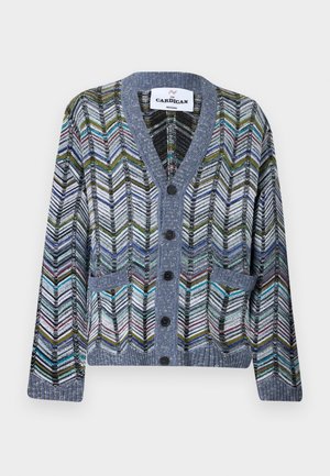 Homme portant un cardigan multicolore à motif chevrons sur une chemise blanche, un pantalon sombre et des baskets écrues, debout devant un fond uni.
