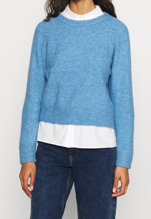 Personne portant un pull en tricot bleu clair superposé à une chemise blanche boutonnée et un jean bleu foncé, debout devant un fond uni.