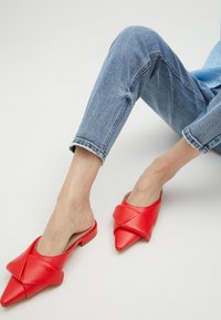 Crvene kožne slip-on mule s šiljastim vrhom i uvijenim detaljem. Nosi se s zarubljenim svijetloplavim trapericama na neutralnoj pozadini.