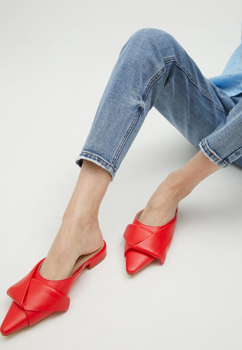 Crvene kožne slip-on mule s šiljastim vrhom i uvijenim detaljem. Nosi se s zarubljenim svijetloplavim trapericama na neutralnoj pozadini.